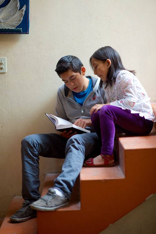 A young man in jeans and a pullover sits on the steps while his little sister, in purple pants and a flowered shirt, sits on a step above him and reads along with him in the Book of Mormon.