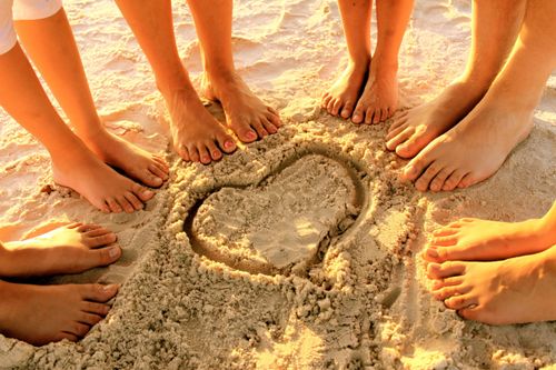 Six pairs of feet are seen standing in a circle around a heart drawn in the sand, with the sun shining in the background.