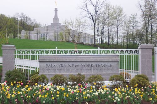 The granite sign outside of the Palmyra New York Temple fence, with the temple seen in the background beyond the green lawns.