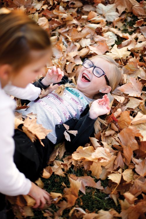 A young girl, out of focus, is outside on an autumn day, piling leaves on her younger sister, who is in focus.