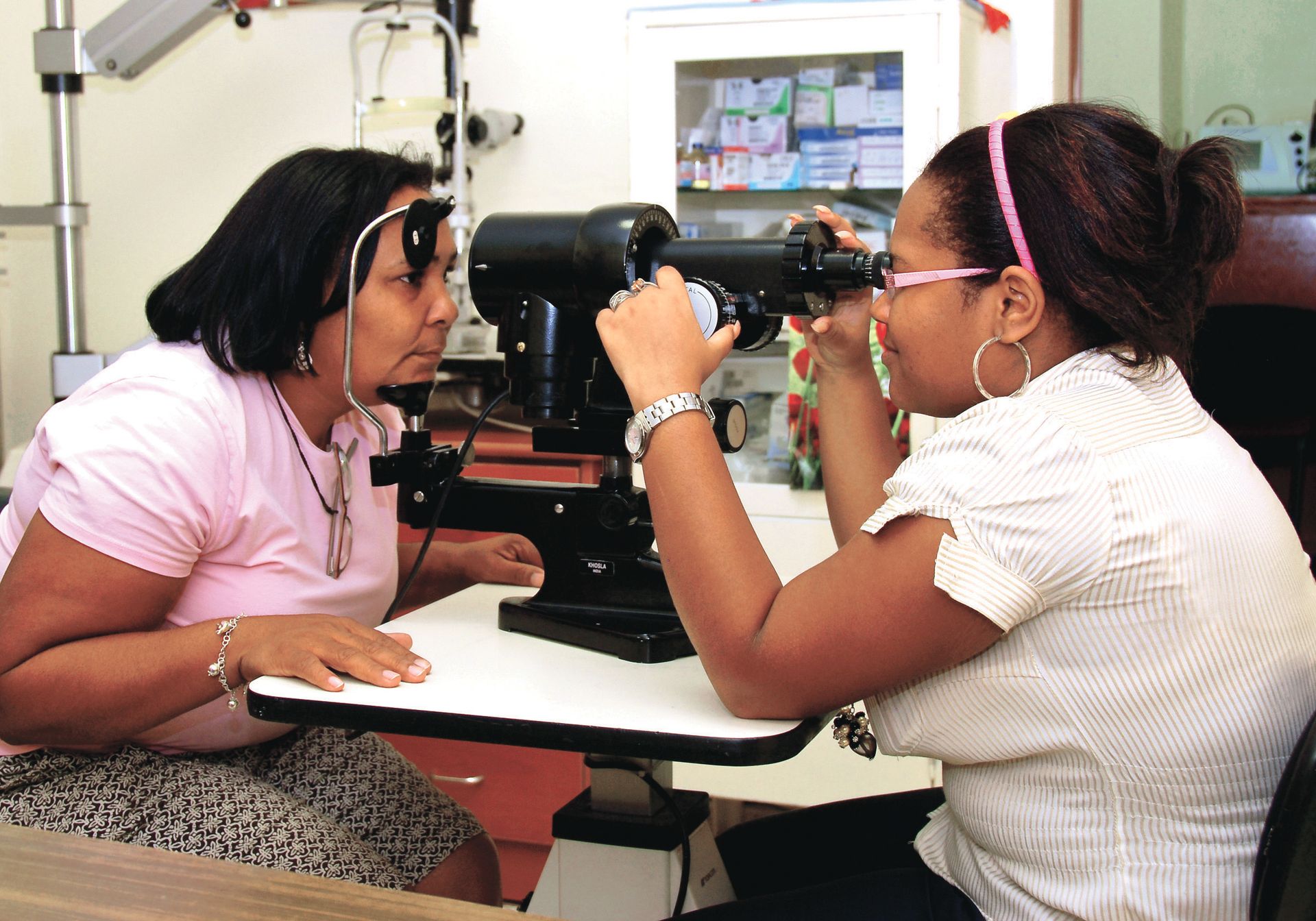 A Dominican woman sitting and resting her chin on an eye examination device while a doctor checks her eyesight.