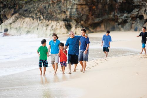A mother and father hold hands with their three sons and walk in the sand along the beach.