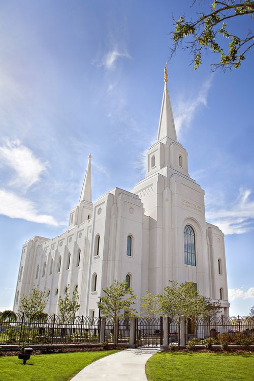 The Brigham City Utah Temple, grounds, and fence on a sunny day, with a blue sky and white clouds.