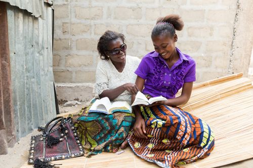 A mother sits next to her daughter on a mat, and they hold their scriptures open on their laps.