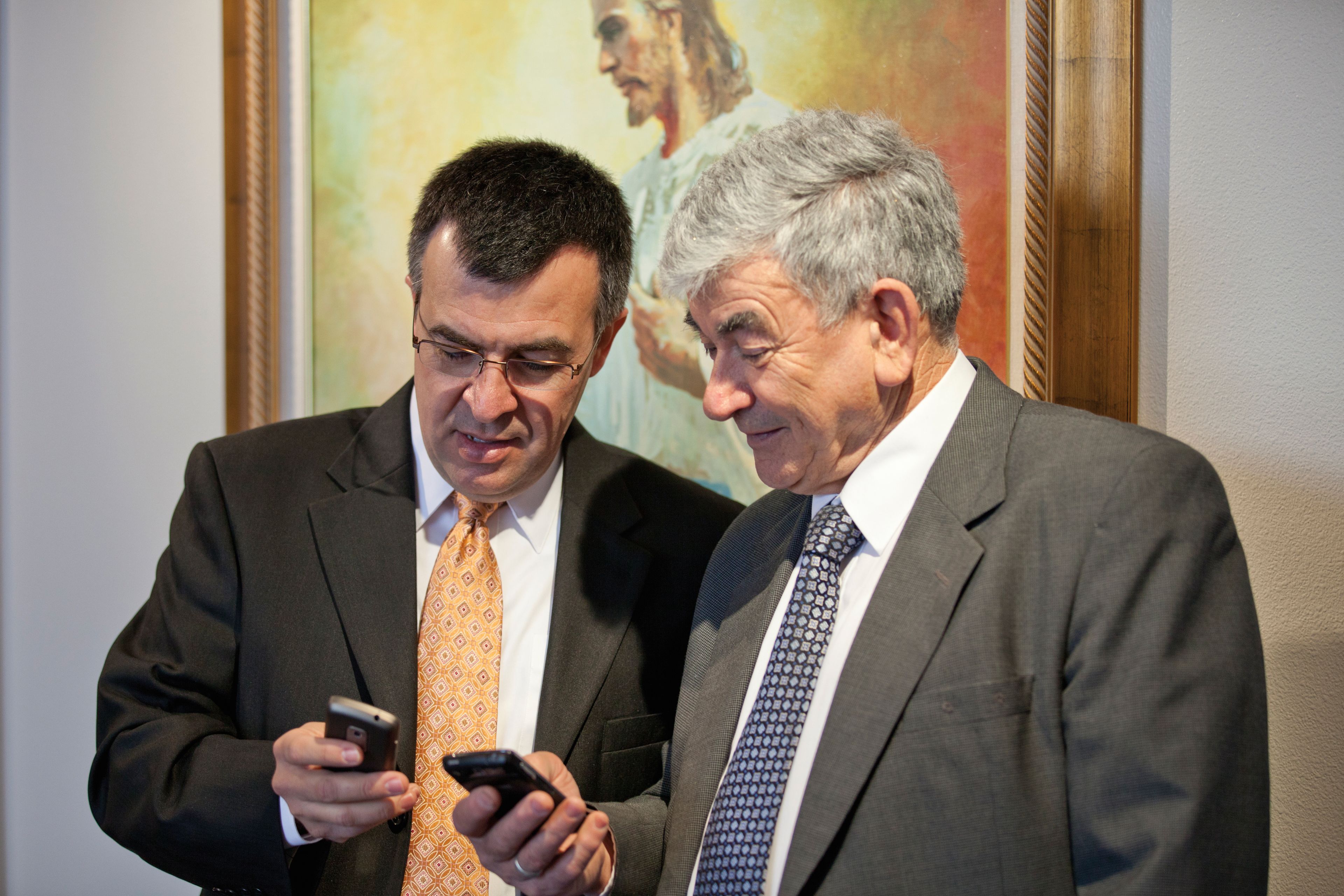 Two older men stand in a meetinghouse and look at the smartphones in their hands.