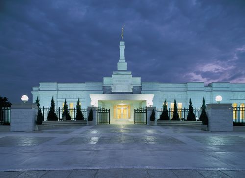 The Oklahoma City Oklahoma Temple entrance at night, with the lights on outside of the temple and warm light coming out from the windows and doors.