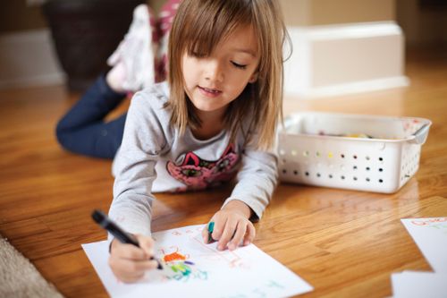 A young girl with straight brown hair lies on the floor and colors with markers on a piece of paper.