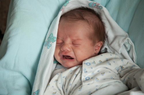 A newborn wrapped in a blanket and lying on a blue pillow, crying.