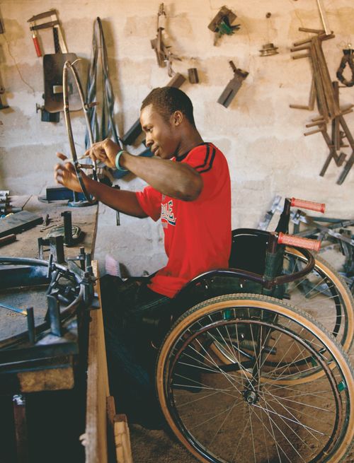 A man from Africa in a bright red shirt, sitting in a wheelchair and repairing a round wheel piece from another wheelchair.