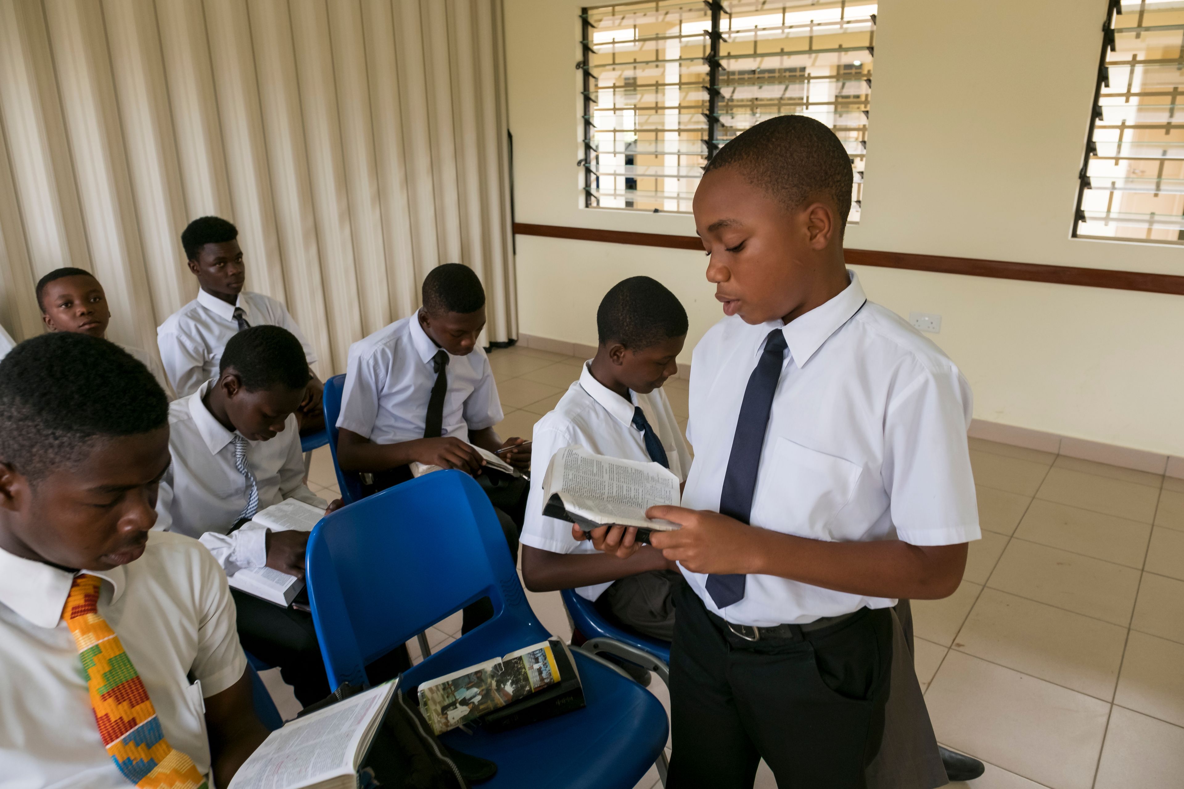 A group of young men sitting in a priesthood class in Ghana. One young man is standing and reading from the scriptures.