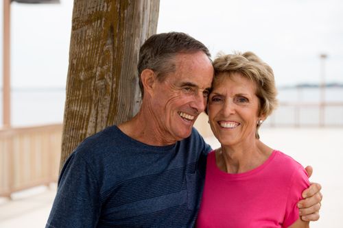 A portrait of an elderly man in a blue shirt hugging his wife, who is wearing a pink shirt, as they stand outside together.