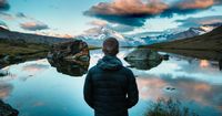 A man stands at the edge of an Alpine lake high in the Swiss Alps. There are mountain peaks and clouds in the distance.