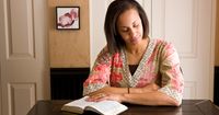 A woman sitting at a table in her home.  She has her eyes closed in prayer.  An open book of scripture is on the table in front of her.