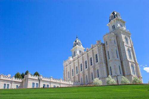 A side view of the Manti Utah Temple on top of a green grassy hill with blossom-covered trees and a blue sky in the distance.