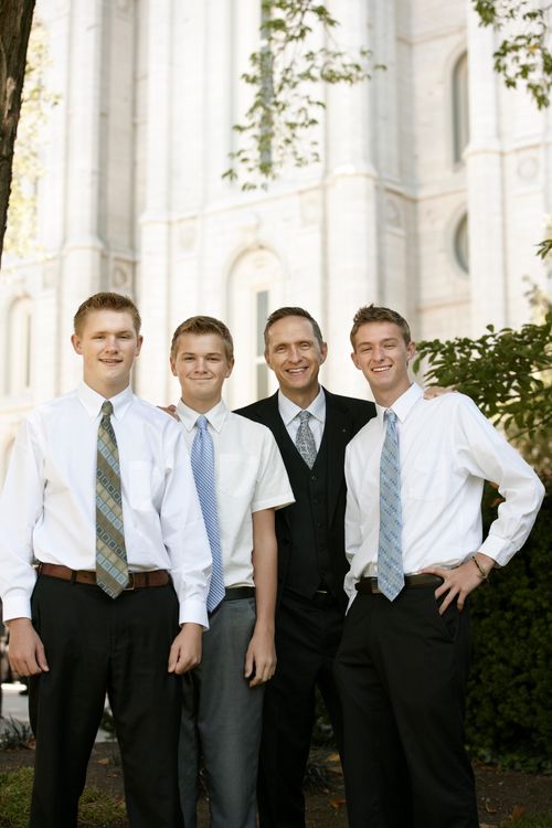 A man in a black suit standing beside three young men in white shirts and ties outside the temple, waiting for a session of general conference.