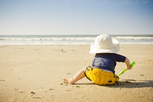 A baby in a sun hat, yellow shorts, and blue shirt plays in the sand with a plastic shovel on the beach.