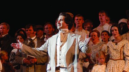 A man dressed in pioneer era clothing stands on a stage in front of a large group of performers also dressed in pioneer clothing. 