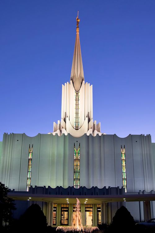 The front of the Jordan River Utah Temple at night, with the lights on inside and outside the temple, illuminating the building and the water fountain in front.