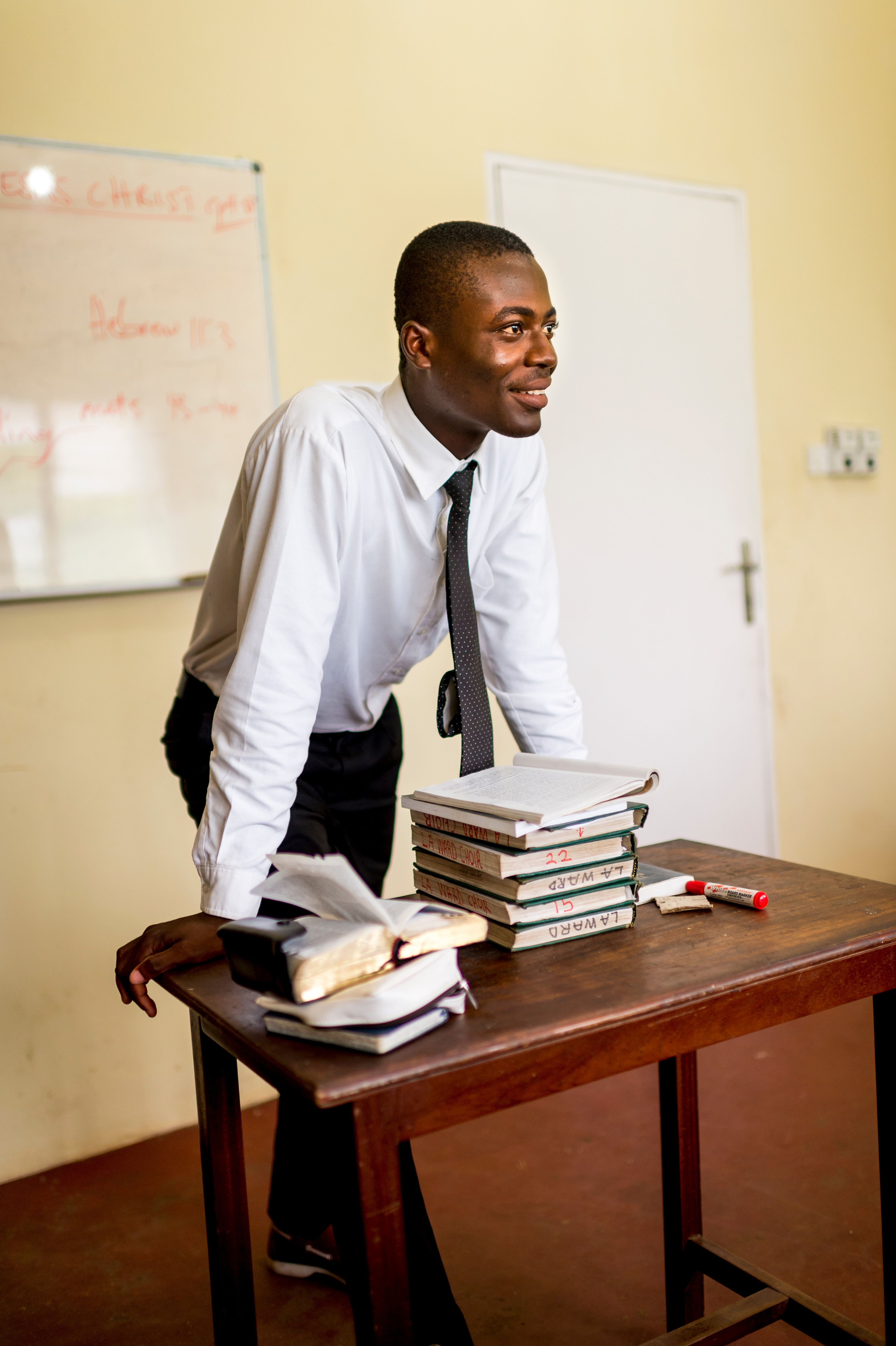 A teacher stands at the front of his classroom with hymnbooks and scriptures on a table in front of him.  