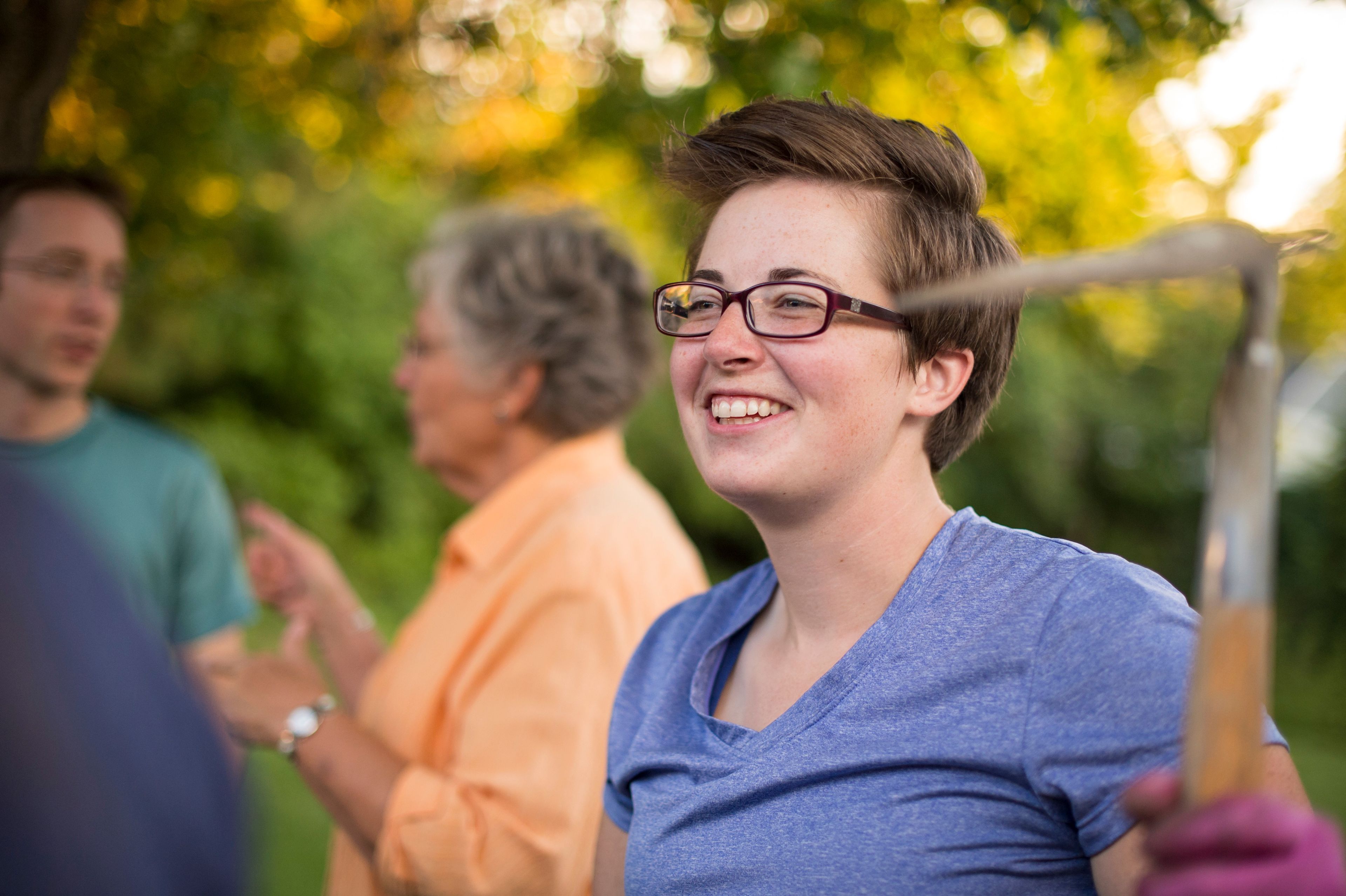 A young woman smiles and holds up a garden tool while doing yard work.  