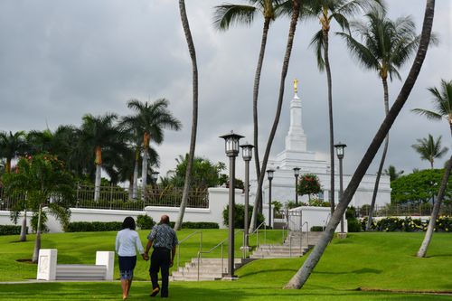 A woman in a white blouse and blue skirt holding hands with her husband in a blue shirt and pants as they walk on the Kona Hawaii Temple grounds.