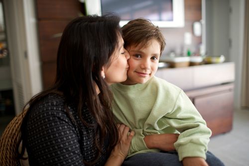 A dark-haired woman kisses her son on the cheek while he looks out at the camera.
