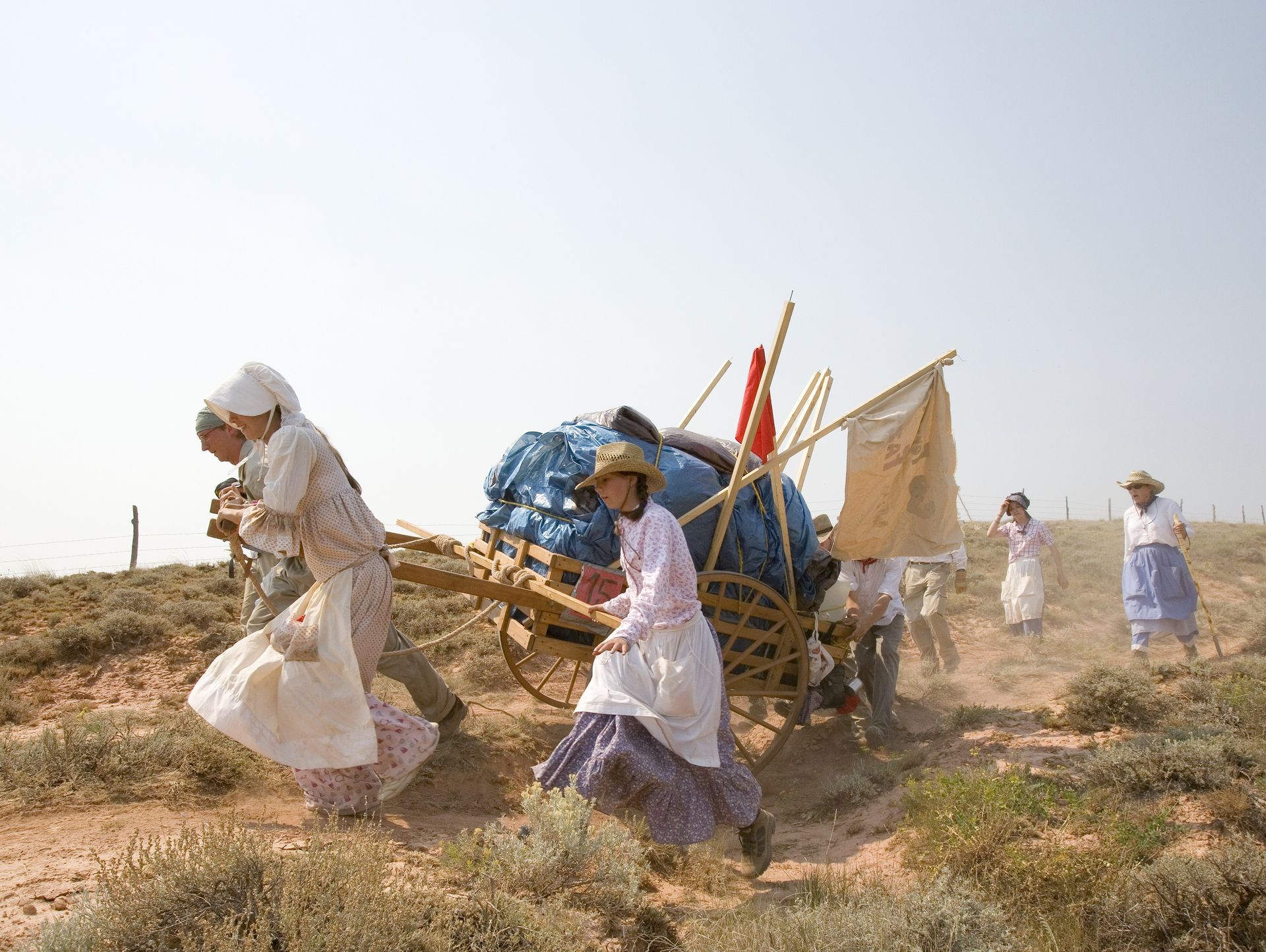 A group of women dressed as pioneers pull a handcart together over a rocky path.