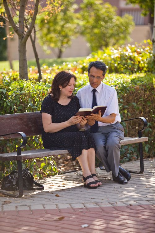 A man in a white shirt, black tie, and gray pants sits on a wooden bench next to a woman in a black dress and sandals as they read the scriptures together.
