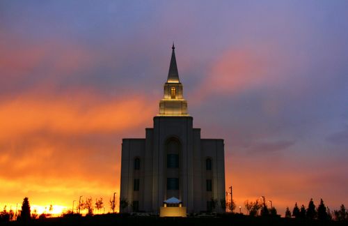 A front view of the Kansas City Missouri Temple at sunset, with orange and red colors in the sky and the lights on the temple illuminating the stone.