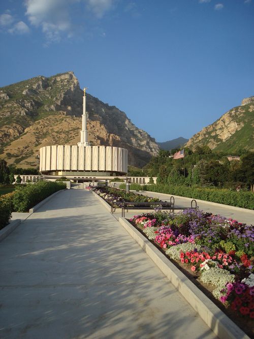 A tree-lined path leading to the Provo Utah Temple on a sunny day, with a tall mountain seen in the background behind the temple.