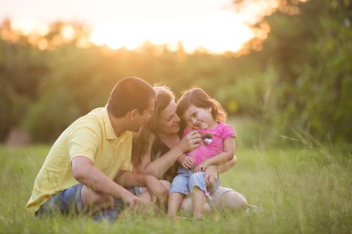 A mother, father and young girl sitting on a lawn in bright sunlight.  They are laughing.