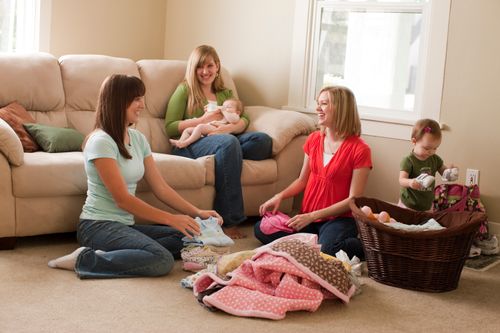 Young adult women helping a young mother fold her laundry.  The mother is holding a baby.
