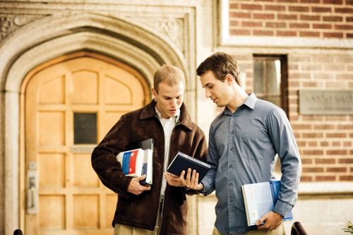 A male college student in a blue shirt standing while looking at the Book of Mormon with another male college student in a brown jacket.