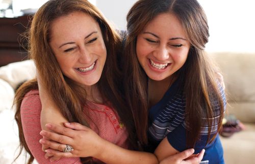 Two young women with long hair, both wearing striped shirts, hug one another and laugh together.