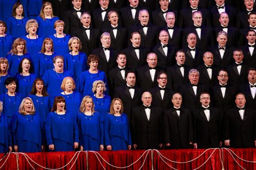 Rows of women in blue dresses and men in black tuxedos singing in the Mormon Tabernacle Choir behind a red partition.