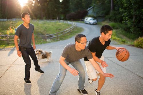 A father plays basketball with his two teenage sons in their driveway, with their dog running behind them.