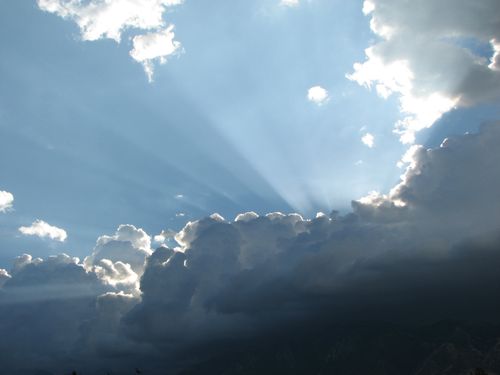 Sun rays extend upward above a group of dark gray clouds into the blue sky.