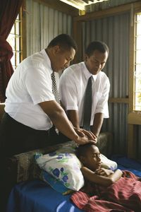 Two Fijian men administering to a young girl lying in a bed.