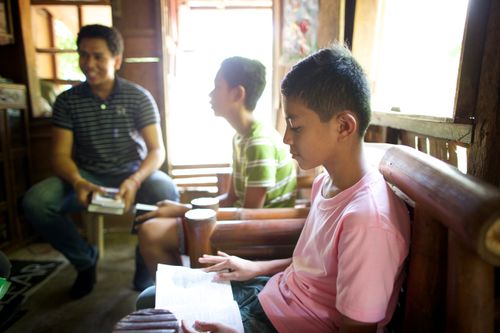 Two ministering brothers in a home, sitting near a young man in a pink T-shirt and reading from the scriptures, with the sun lighting the room.