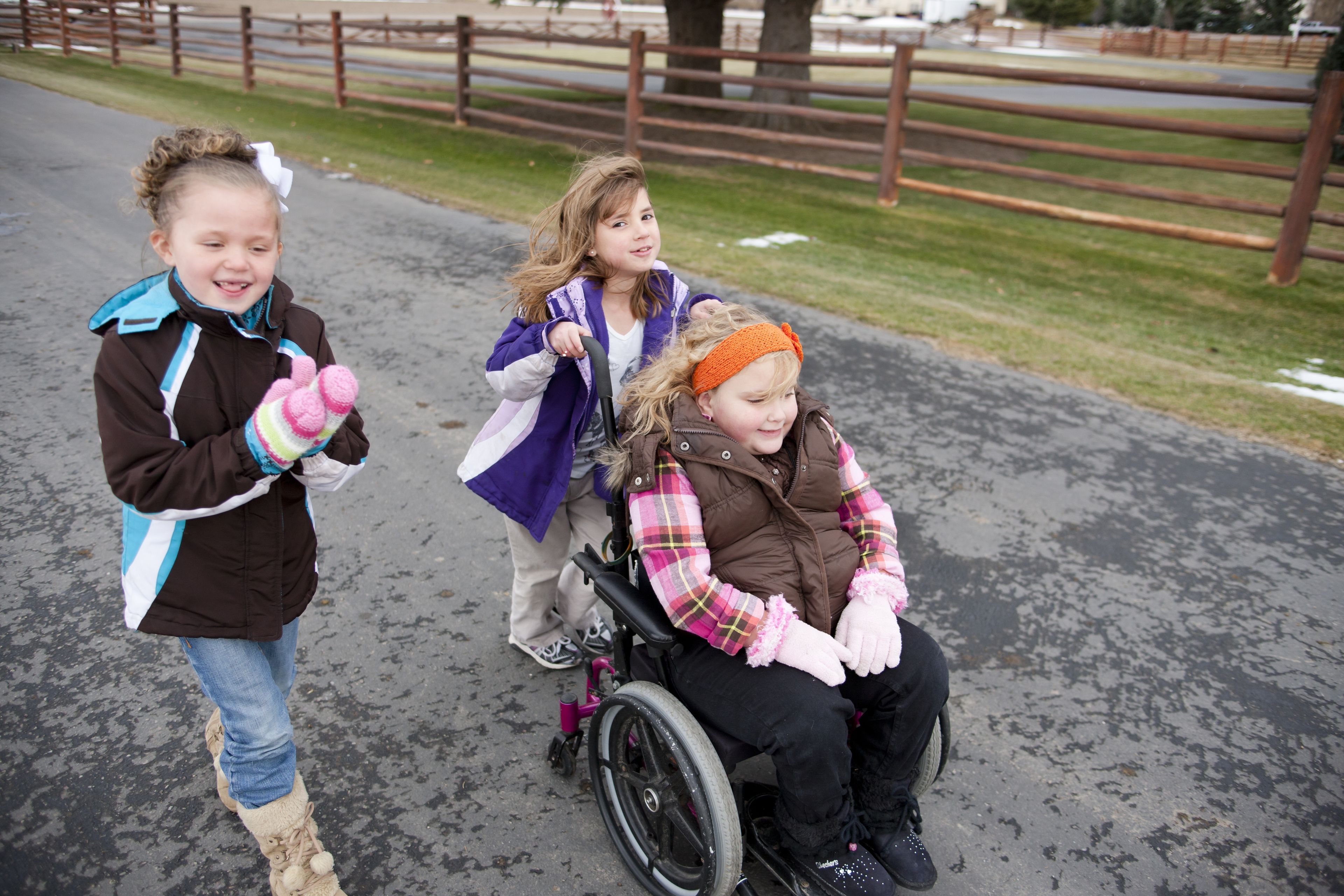 Two girls walk together while one pushes a third girl in a wheelchair.