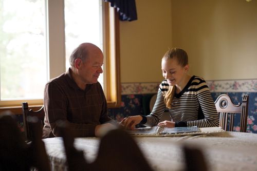 An elderly man and his granddaughter sit together at the kitchen table to look at a family photograph album.