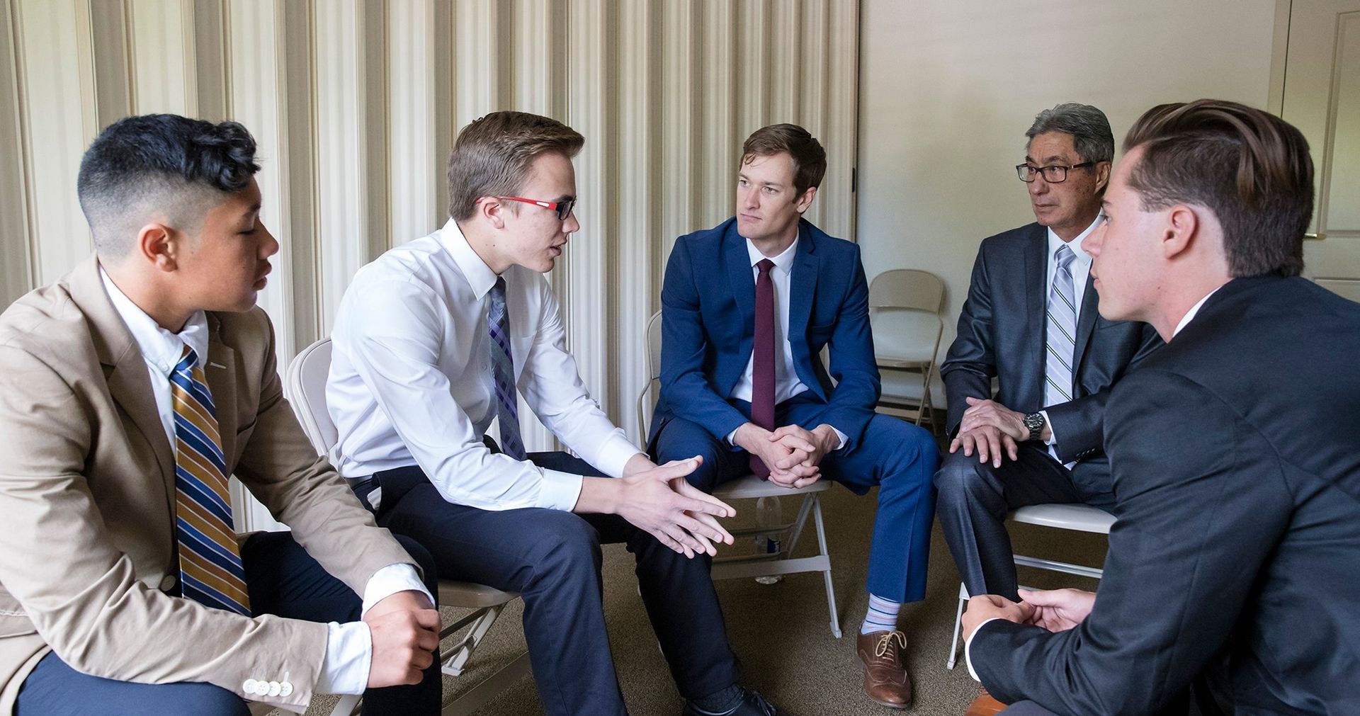 Three men are together in a church building. They appear to be in a classroom of an LDS Meetinghouse. Two of the men appear to be leaders and the other a youth. They are talking with one another, having a meeting.