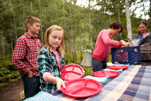 A young girls sets red plates on a picnic table with her mother and siblings at a campground.