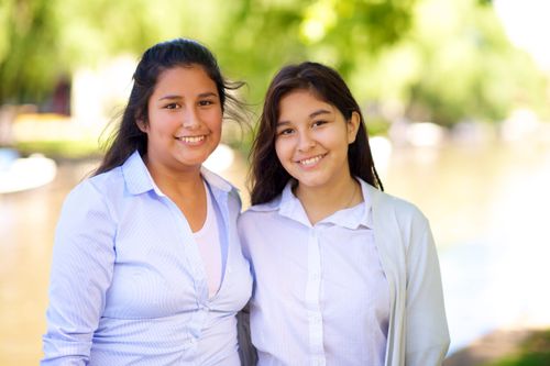 A portrait of two young sisters from Argentina, standing side by side outdoors and smiling. They are wearing light-colored shirts.