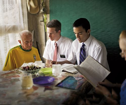 Two elder missionaries in Bulgaria sitting next to an elderly man in his home as they read scriptures together.