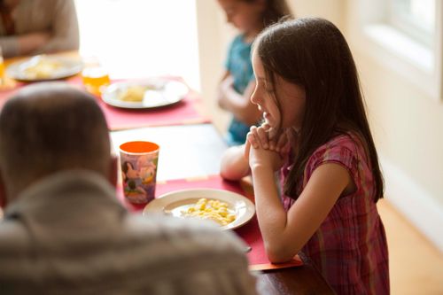 A little girl clasps her hands and leads her family in prayer before they eat dinner at the table.