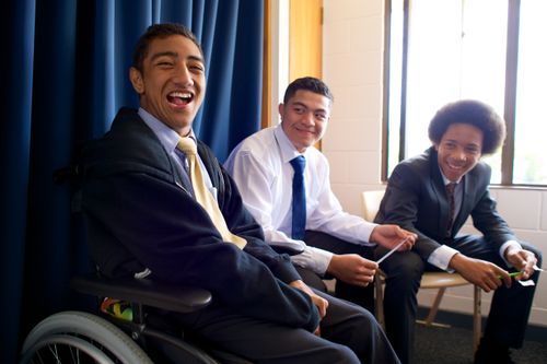 One young man in a suit and tie sits in a wheelchair, with two other young men sitting next to him and smiling.