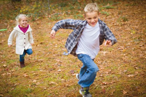 A boy running with outstretched arms ahead of his younger sister over a leaf-covered ground.
