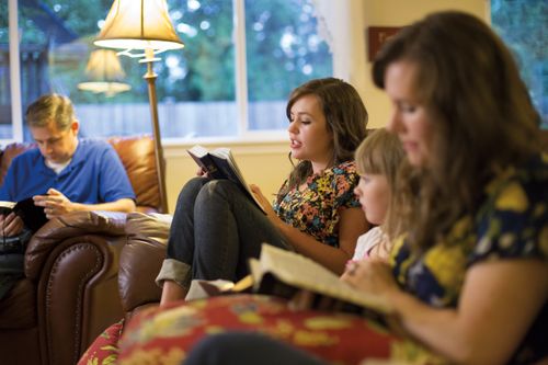 A young woman reads from the scriptures while her mother, father, and sister sit in the living room with her and follow along.