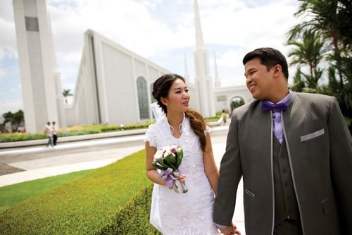 A bride in a lace dress with a bouquet of flowers holds hands with the groom in a gray suit as they walk outside of the Manila Philippines Temple.